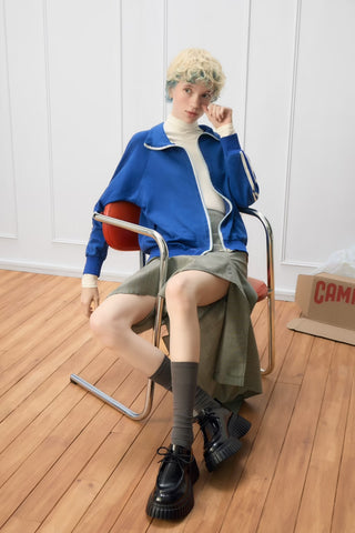 Side view of a woman seated on a chair wearing Camper Pix BCN black leather shoes, styled with a blue jacket and green skirt in a white-walled room with wooden floors.